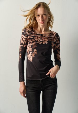 Mujer rubia con camiseta negra de manga larga con patrón floral marrón y vaqueros negros, posando con una mano cerca del bolsillo sobre un fondo sencillo.