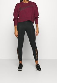 Žena oblečená v tmavě burgundském svetru Nike a černých legínách s žebrovanými výřezy u kotníků, stojící na bílém pozadí.