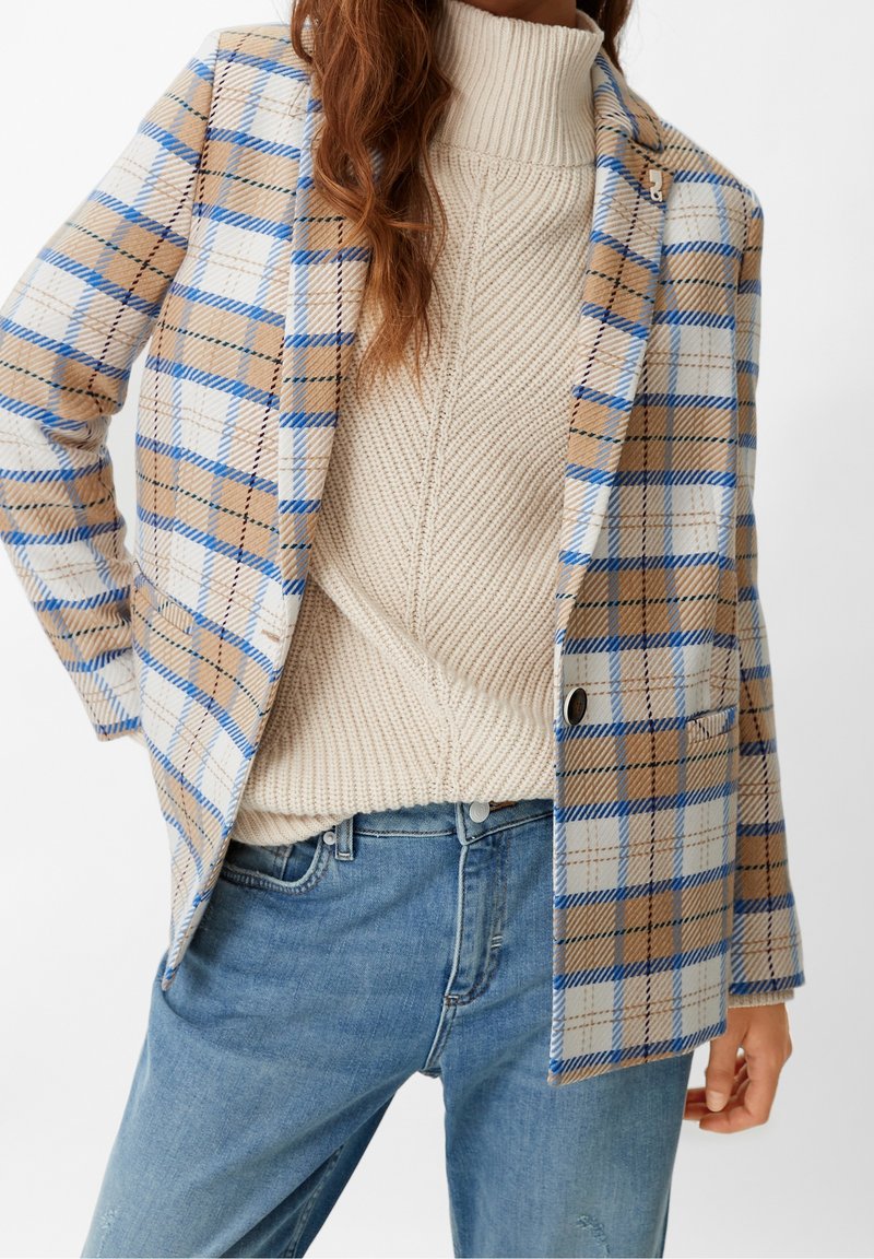 Femme portant un pull à col roulé en maille beige, un blazer à carreaux bleu et beige, et un jean bleu, une main sur la hanche.