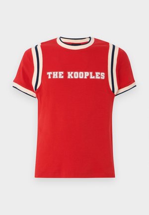 Henkilö, jolla on punainen lyhythihainen paita, jossa lukee "THE KOOPLES", tummat suorat farkut, valkoiset sukat ja mustat loafersit, seisoo paikoillaan.