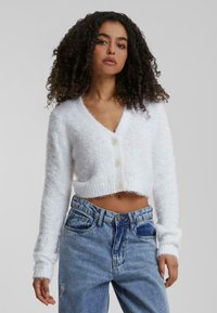 Cardigan court en peluche blanc avec un col en V et cinq boutons, doté de manches longues et d'une texture douce, associé à un jean en denim taille haute.