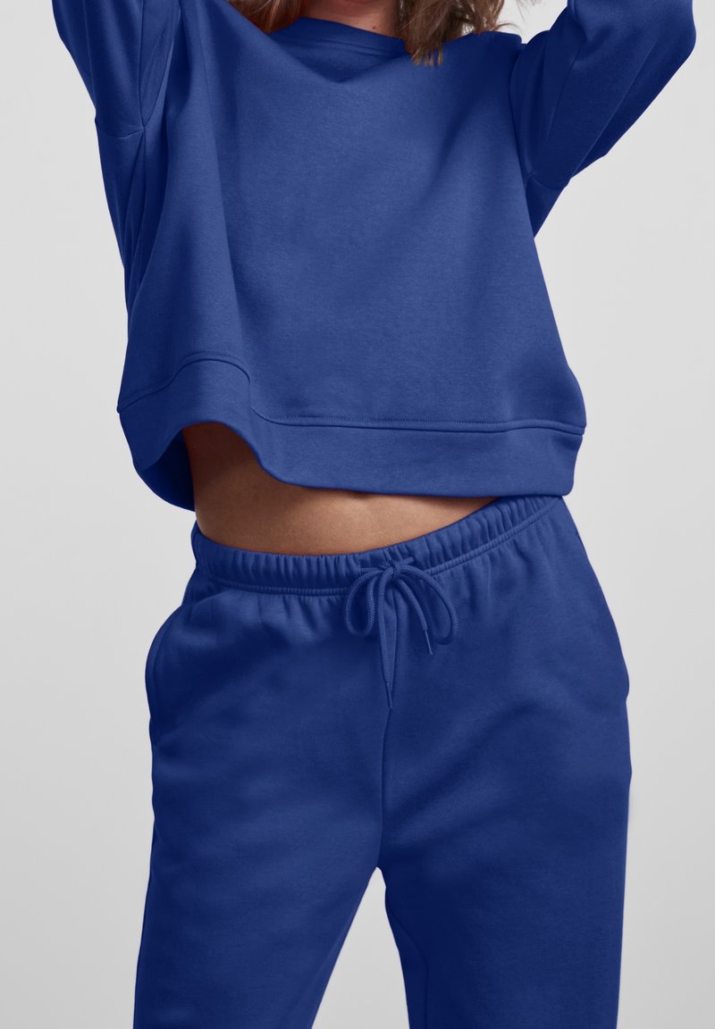 Personne portant un sweat-shirt bleu assorti et un pantalon de survêtement à cordon, levant légèrement les bras, montrant le ventre sur un fond uni.