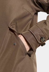 Braune wasserdichte Jacke mit glatter Textur, auffälligen Knöpfen und gesticktem Logo. Verfügt über Seitentaschen und umgeschlagene Manschetten.