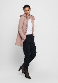 Cappotto lungo rosa chiaro con cappuccio, abbottonato nella parte frontale, sovrapposto a una camicia bianca. Pantaloni cargo neri e stivali con tacco neri completano il look.