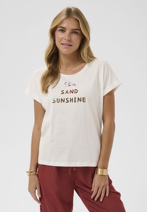 Femme blonde portant un t-shirt blanc avec le texte "Sea Sand Sunshine" et un pantalon rouge, posant avec un bracelet en or et des boucles d'oreilles créoles.