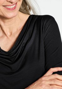 Schwarzes Oberteil mit drapiertem Ausschnitt, aus weichem, glattem Stoff. Lange Ärmel und eine figurnahe Silhouette, die einen subtilen Glanz zeigt.