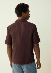 Short-sleeve brown shirt with textured fabric, a small collar, and a straight hem, paired with light blue denim jeans.