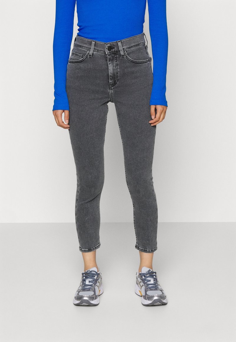 Jeans de tiro alto, ajustados, de color negro desvaído con cinco bolsillos y costuras en contraste, combinados con una camiseta de manga larga azul y zapatillas grises.