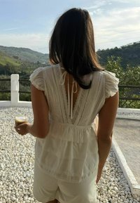 Femme aux longs cheveux bruns portant un haut en dentelle crème et un short, tenant un verre de café, debout sur des galets blancs avec des collines et de la verdure en arrière-plan.