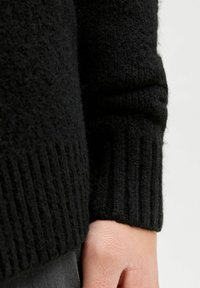 Juodas megztinis su tekstūruotu paviršiumi ir supjaustytais rankogaliais, pagamintas iš minkštos, pūkuotos medžiagos, suteikiančios šilumą.