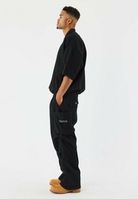 Camicione nero oversize con spalle basse, abbinato a pantaloni cargo neri con tasca a toppa. Calzature in tono sabbia con suola texturizzata.