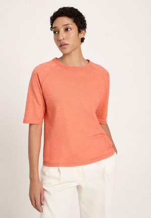 Person with short curly hair wearing a salmon-colored short-sleeve top and white pants, standing against a plain light background.