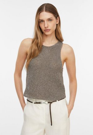 Młoda kobieta z długimi jasnobrązowymi włosami nosi bezrękawnik w kolorze szarym i białe spodnie z cienkim brązowym paskiem, ręce w kieszeniach.