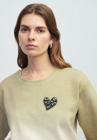 Sweatshirt vert olive avec un col arrondi et des manches courtes, présentant un appliqué en forme de cœur orné de bijoux sur la poitrine.