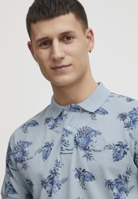 Kortärmad polo t-shirt i ljusblått bomull, med ett blommönster i mörkblått, och med en klassisk krage och knapplist.