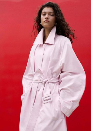 Vrouw met krullend haar die een oversized lichtroze mantel met ceintuur draagt, staand tegen een felrode achtergrond, handen in de zakken.