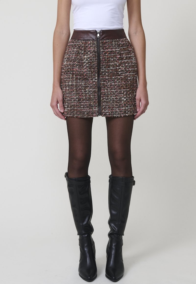 Femme portant une mini-jupe en tweed multicolore avec fermeture éclair devant, des collants noirs transparents, des bottes noires montantes et un haut blanc sans manches.