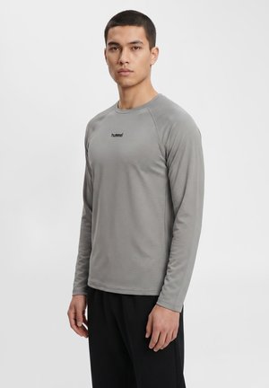 Jeune homme debout face à l'avant, portant une chemise grise unie à manches longues et un pantalon noir sur un fond clair.