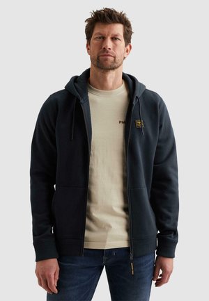 Mann trägt einen marineblauen Kapuzenpullover mit Reißverschluss über einem beigen T-Shirt und blauen Jeans, steht vor einem schlichten hellgrauen Hintergrund.