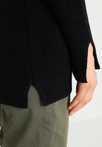 Maglione nero a maglia con texture a coste, dotato di spacchi laterali all'orlo. Abbinato a pantaloni verde oliva chiaro, mano visibile di lato.