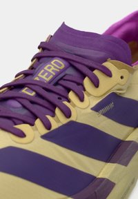 Chaussure de sport violette et beige avec des lacets assortis. Présente une tige texturée, un nom de marque bien visible et un design rayé sur le côté.