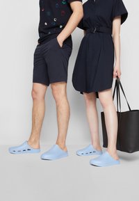 Sapatos azul claros, tipo slip-on, feitos de borracha e com buracos de ventilação, usados por duas pessoas. Um tem uma bolsa preta e os calções do outro são azul-marinho.