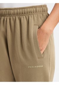 Beige Jogginghose mit elastischem Bund, Seitentaschen und auf dem Oberschenkel in hellerem Beige aufgedrucktem "PEGADOR"-Logo. Weiche Stoffstruktur.
