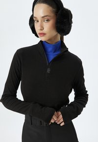 Zwarte ritspullover met lange mouwen en duimgaten, gedragen over een blauwe coltrui, gecombineerd met zwarte broek met riem.