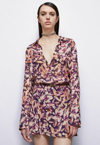 Abito floreale con bottoni in tonalità di viola, arancione e crema. Maniche lunghe con polsini a campana, vita cintrata con una cintura marrone, tessuto morbido.