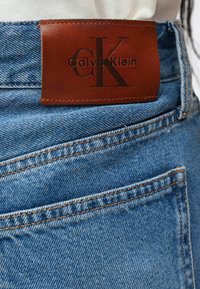 Καφέ δερμάτινη ετικέτα Calvin Klein ραμμένη στην τσέπη μπλε τζιν με ορατά πορτοκαλί ράμματα και λευκό ύφασμα πάνω από τη μέση.