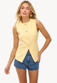Mouwloze gele vest met een gestructureerd ontwerp, vier knopen aan de voorkant en zijzakken, gecombineerd met denim shorts. Zachte textuur.