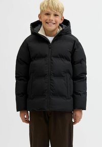 Smilende blond dreng iført sort puffjakke med hætte, beige inderskæring, hvid skjorte og brune bukser foran en ensfarvet baggrund.
