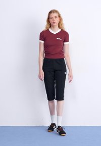 Rotes T-Shirt mit weißem Saum, kombiniert mit schwarzen knielangen Hosen mit weißen Seitstreifen. Das Model trägt schwarze Sneakers und weiße Socken.
