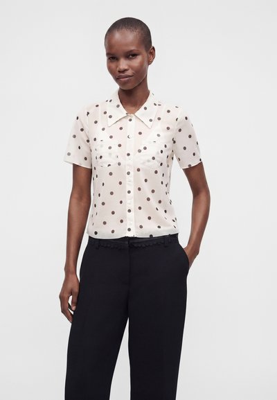 Femme portant un chemisier blanc à manches courtes avec des pois marron et un pantalon noir, debout devant un fond clair uni.
