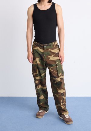 Homme portant un débardeur noir, un pantalon cargo camouflage et des baskets marron debout sur un sol bleu contre un mur blanc.