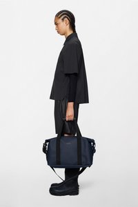 Μαύρη αδιάβροχη τσάντα duffel με ανθεκτικούς μαύρους ιμάντες, ορθογώνιο σχήμα, διακριτικό λογότυπο και λεία υφή, κρατημένη από ένα άτομο με μαύρη ενδυμασία.