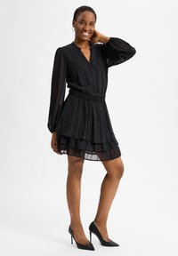 Schwarzes Kleid mit langen Ärmeln, gefalteten Stufen am Saum, V-Ausschnitt und strukturiertem Stoff. Getragen mit schwarzen High-Heels.