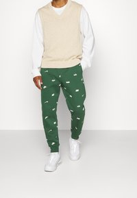 Beige stickad väst över en vit skjorta med långa ärmar, ihop med gröna mönstrade joggers med vita logotyper, kompletterat med vita sneakers.