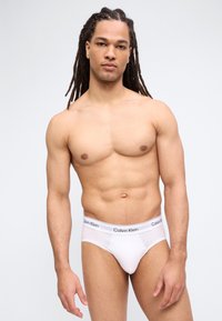 Μυώδης άνδρας με μακριά κοτσίδα dreadlocks, φοράει λευκά εσώρουχα Calvin Klein, σε απλό φωτεινό φόντο.