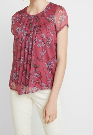 Blusa corta in maroon con motivi floreali blu e verdi. Tessuto trasparente con pieghe sul davanti, abbinata a pantaloni bianchi.