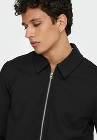 Svart jakke med glidelås og krage, med glatt tekstur, sølvfarget glidelåshardware, og en tettsittende design.