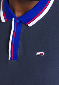 Polo marine en tissu texturé, avec un col bleu orné de rayures blanches et rouges, et un petit logo brodé sur la poitrine.