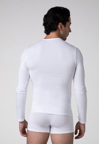 Camicia bianca a maniche lunghe realizzata in un materiale liscio, aderente, con un semplice collo tondo. Indossata con biancheria intima bianca coordinata.
