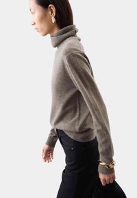 Beige Rollkragenpullover mit geripptem Kragen und Bündchen, kombiniert mit schwarzen Jeans. Auffälliges goldenes Armreifen-Band am linken Handgelenk, mit glatter Textur.