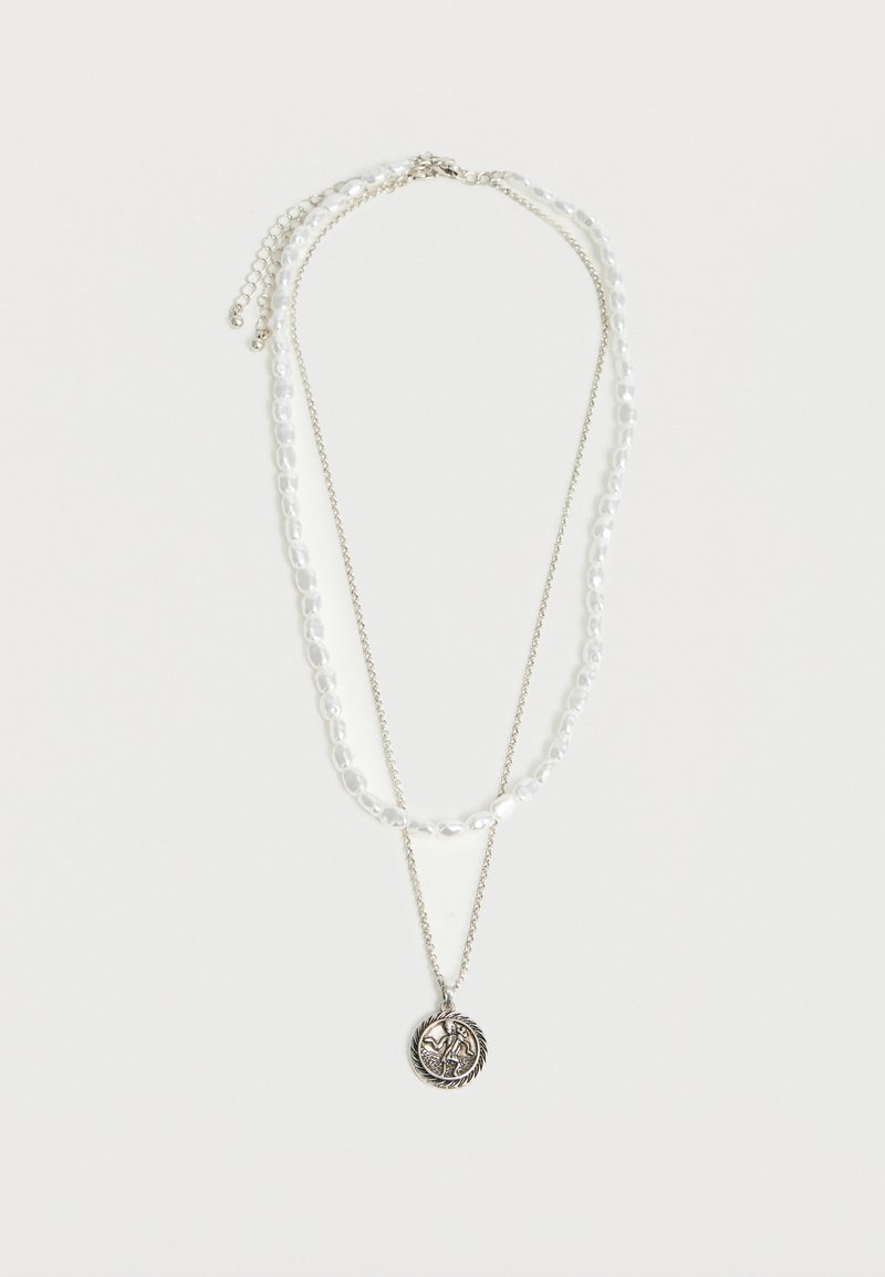 Διπλό κολιέ με μια αλυσίδα από λευκές πέρλες και μια λεπτή ασημένια αλυσίδα που κρατάει ένα στρογγυλό μενταγιόν με ανάγλυφη φιγούρα.