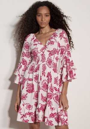 Frau mit lockigem Haar trägt ein weißes Kleid mit rotem Blumenmuster und gerafften Dreiviertelärmeln, steht vor einem schlichten Hintergrund.