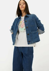 Jeansowa kurtka w ciemnoniebieskim kolorze z zapięciem na guziki, dwoma kieszeniami na piersi i podwiniętymi rękawami, noszona na graficzny t-shirt i luźne dżinsy.