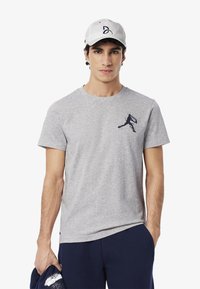 Camiseta de algodón gris con cuello redondo, que lleva un gráfico en azul marino de un jugador de baloncesto en el pecho. Combinada con una gorra blanca.