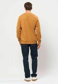 Homme portant un sweat-shirt moutarde, un jean bleu foncé et des baskets blanches, debout, dos à un fond clair et uni.