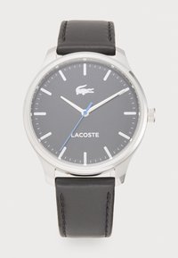 Lacoste laikrodis su sidabriniu nerūdijančio plieno korpusu, pilku ciferblatu, mėlyna antrine rodykle ir juodu odiniu laikrodžio dirželiu su siuvinėjimu.